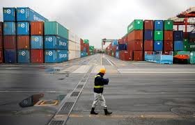 A single worker walks across an empty area of a shipping port in Japan, surrounded by stacks of shipping containers on both sides. The sky is overcast, suggesting a gloomy atmosphere.